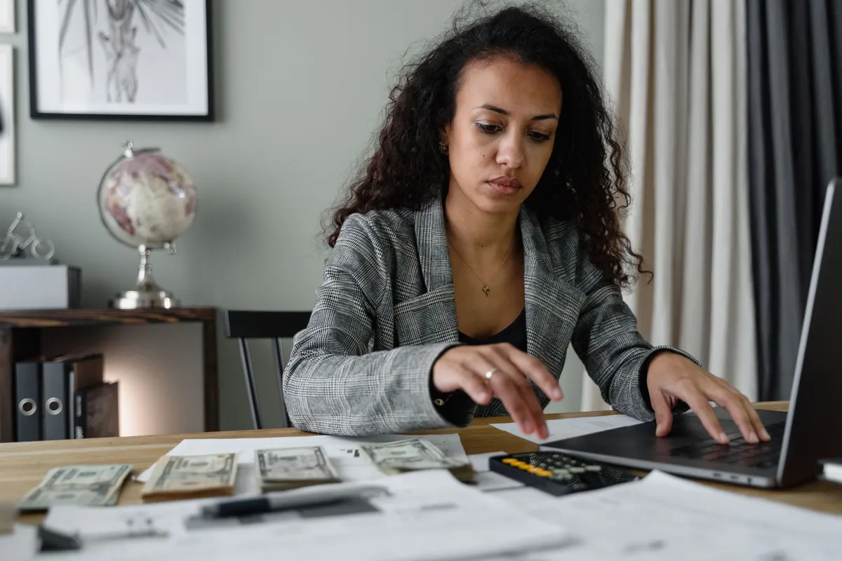 A woman at a desk with a laptop, a calculator and stacks of cash, working through her numbers