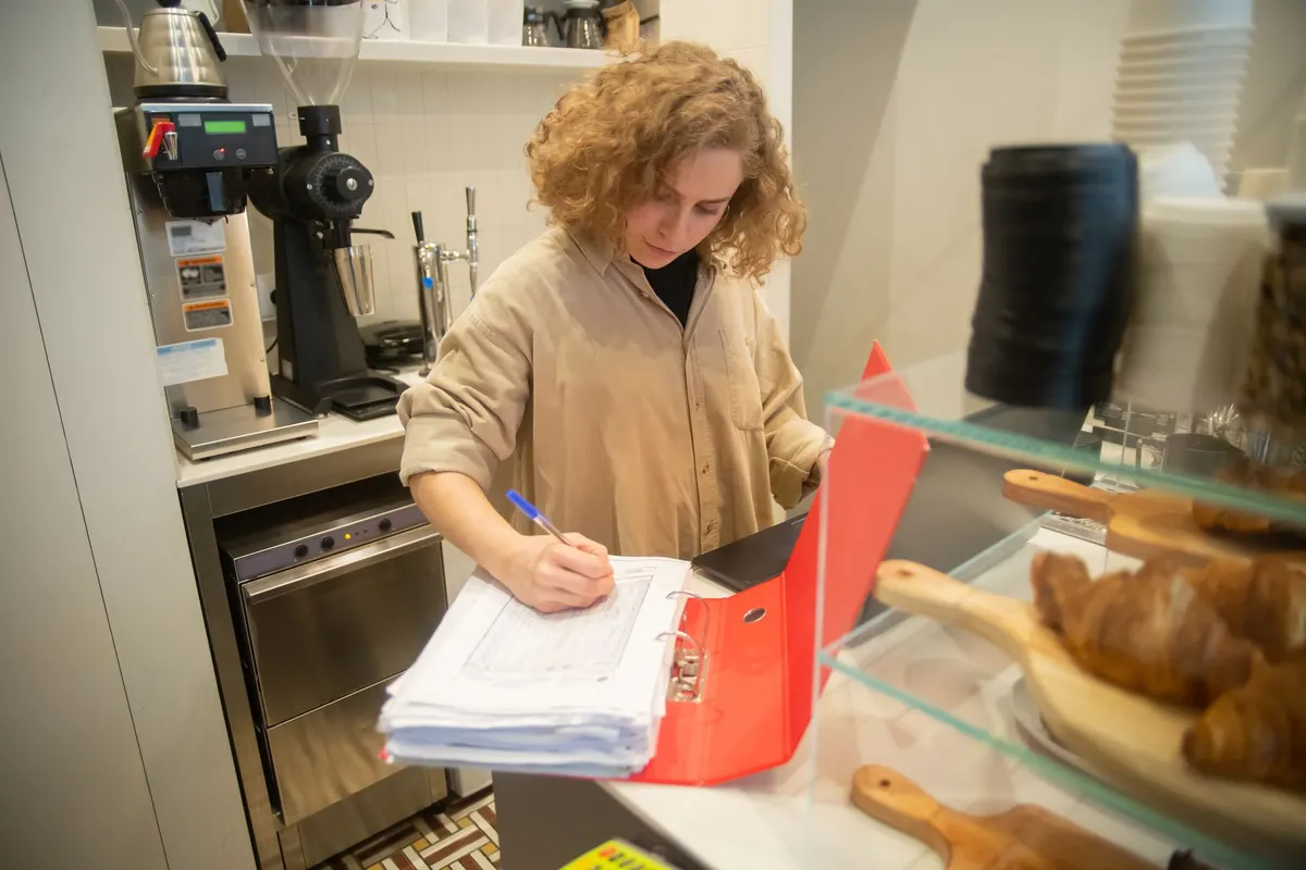 A café owner behind her counter making notes on a thick stack of papers