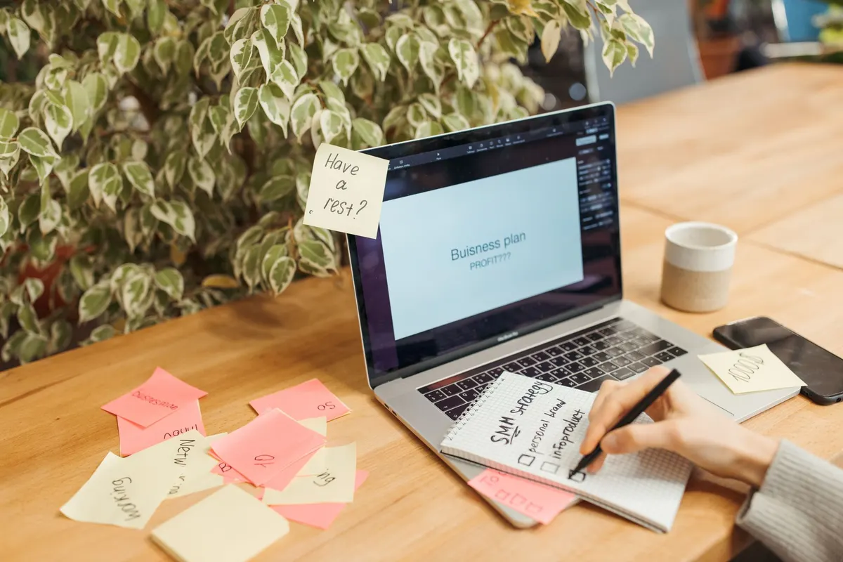 A person writing a plan in a notebook beside a laptop and a spread of sticky notes
