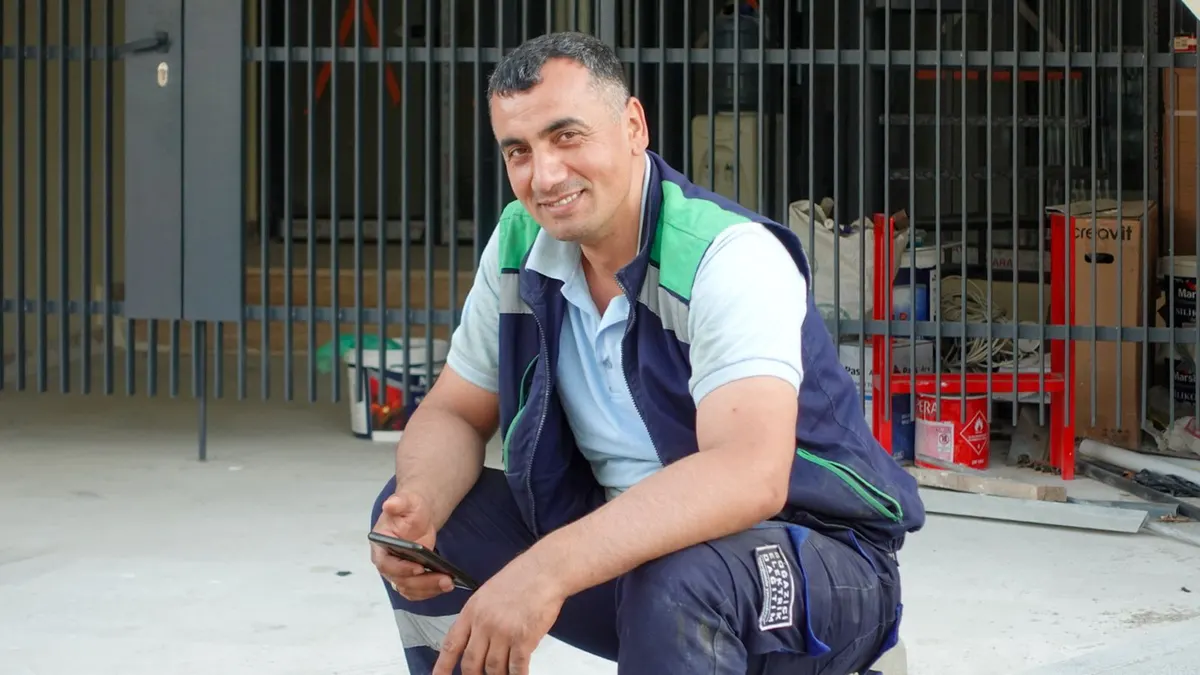 A smiling tradesman in work clothes crouched at a job site, holding his phone