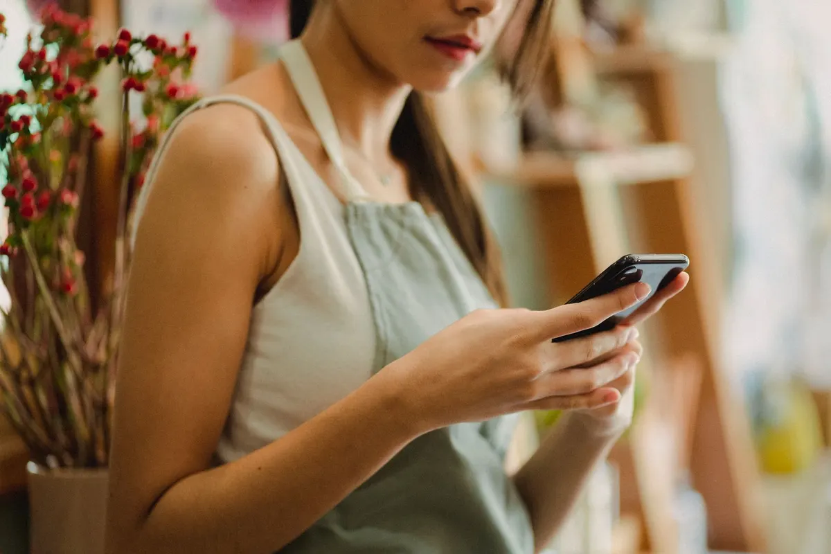 A florist in an apron typing a message on her phone inside her flower shop